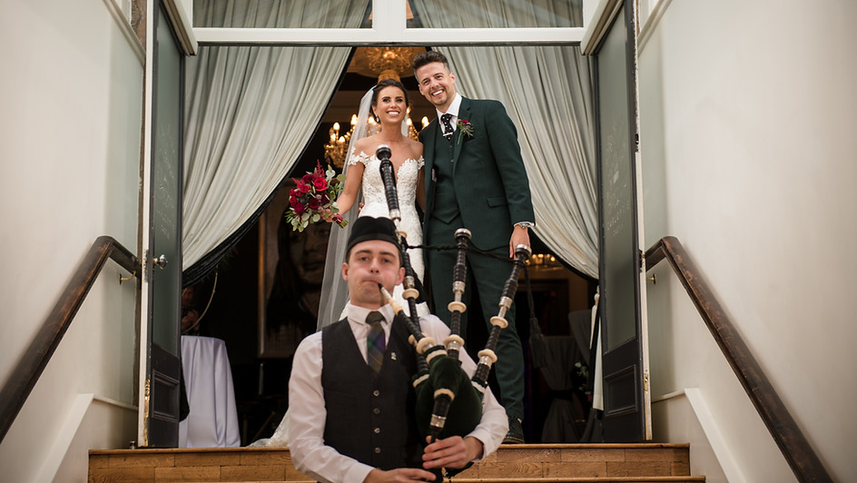 Scottish Piper Hire playing bagpipes whilst marching down stairs in front of a bride and groom inside a reception venue. 