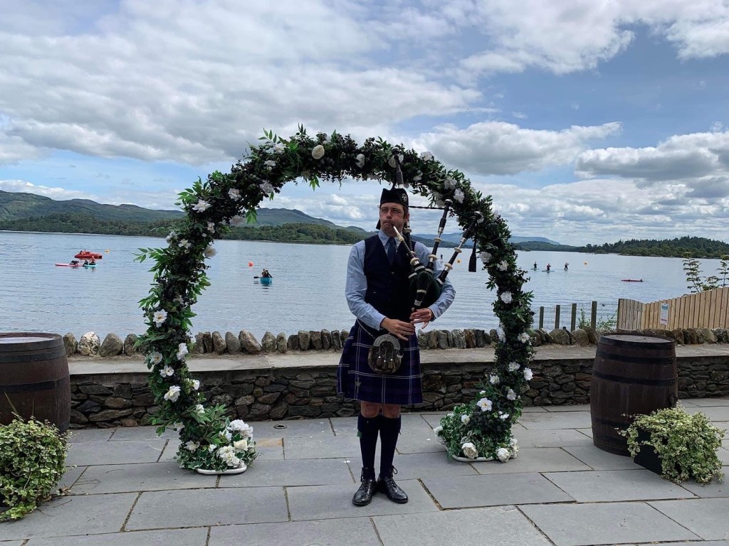 Scottish Piper Hire in traditional highland dress playing bagpipes in front of loch lomond and a flower arch.